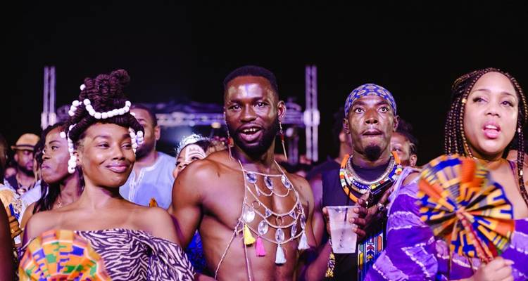 Scène de festival d'Afrobeat nocturne avec des participants vêtus de couleurs vives qui chantent et dansent.