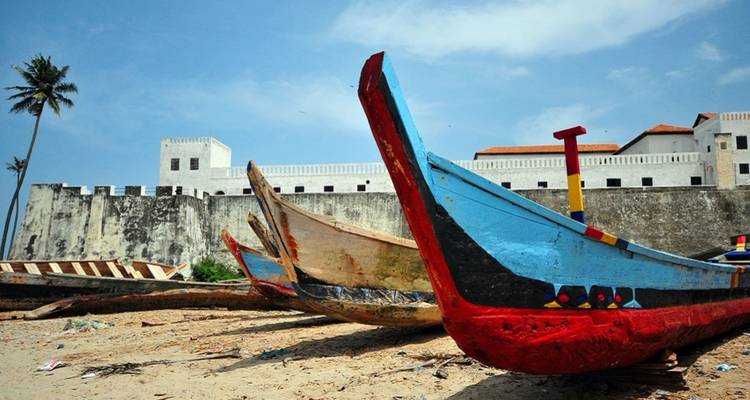 Leuchtend bemalte Holzfischerboote ruhen auf einem Sandstrand, während die weißen Mauern des Elmina Castle dahinter aufragen.