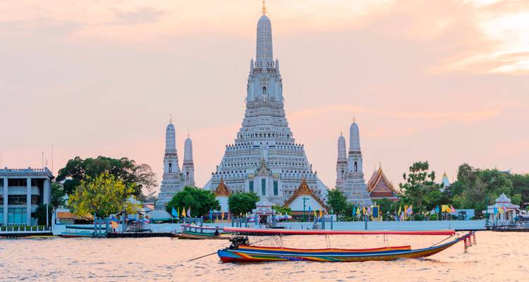 Los elegantes picos del templo Wat Arun brillan con una luz de atardecer pastel junto al río Chao Phraya con una barca de cola larga pasando.
