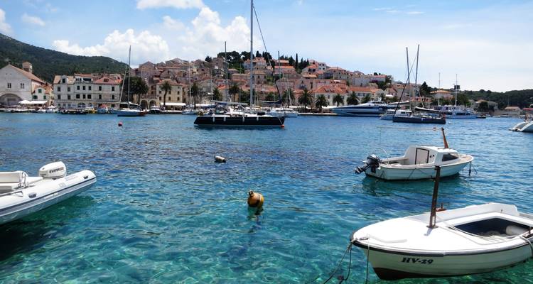 Crystal-clear harbor waters with sailboats anchored before a hillside town of pastel houses and church steeple