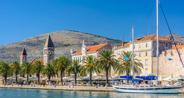 Waterfront promenade of Trogir lined with palm trees, stone towers and tiled rooftops against clear sky