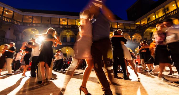 Foule floue dansant le tango sous des lumières chaleureuses dans une cour ouverte la nuit.