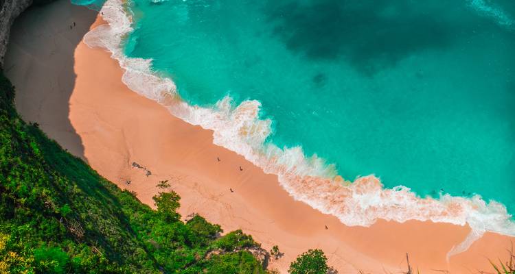 Luchtfoto van een turquoise baai met zacht golfslag die een gebogen perzikkleurig zandstrand ontmoet, omgeven door groene klliffen.
