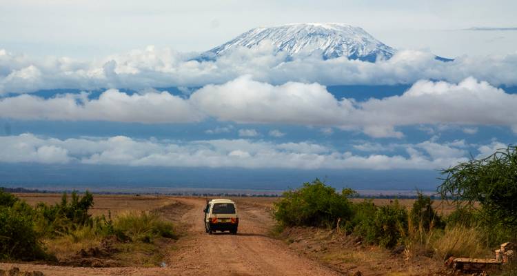 Ein Safari-Geländewagen fährt eine staubige Piste hinunter auf den schneebedeckten Mount Kilimanjaro zu, der über geschichteten Wolken aufragt.