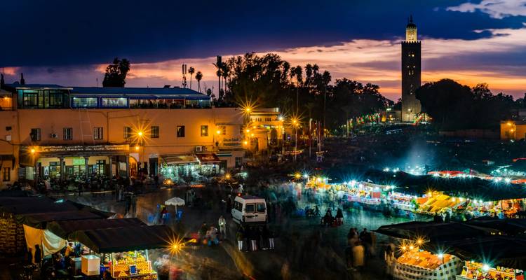 La nuit tombe sur le marché vibrant de la Jemaa el-Fnaa avec ses étals illuminés et la tour de la Mosquée Koutoubia