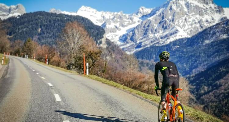 Un ciclista anda en bicicleta por una carretera de montaña con picos nevados que se alzan al fondo.