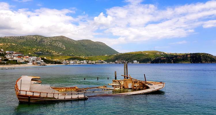 Un navio naufragado de madera envejecida descansa en aguas azules cristalinas cerca de un pequeño pueblo costero albanés.