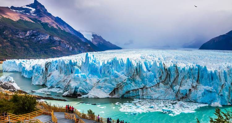 Massieve blauwe Perito Moreno Gletsjerwand met loopbrug uitkijkpunt en bergen erachter.