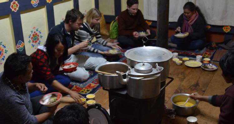 Grupo de viajeros sentados en el piso de madera alrededor de una estufa compartiendo una comida tradicional butanesa.