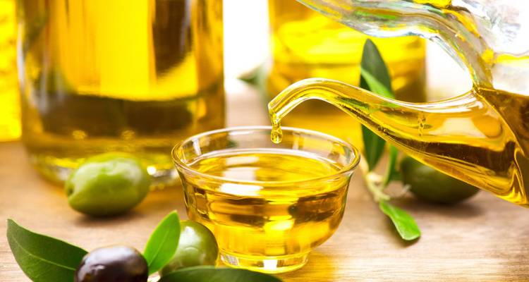 El aceite de oliva dorado se vierte desde una jarra de vidrio en un pequeño cuenco rodeado de aceitunas frescas.