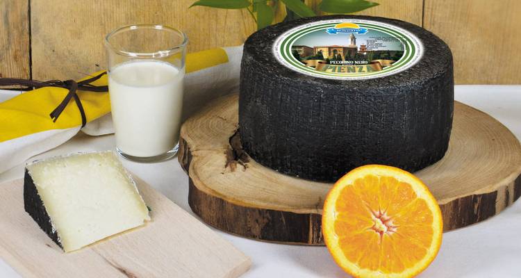 Una rueda de queso Pecorino oscuro con un vaso de leche, una rodaja de naranja y un trozo de queso en una tabla de madera.