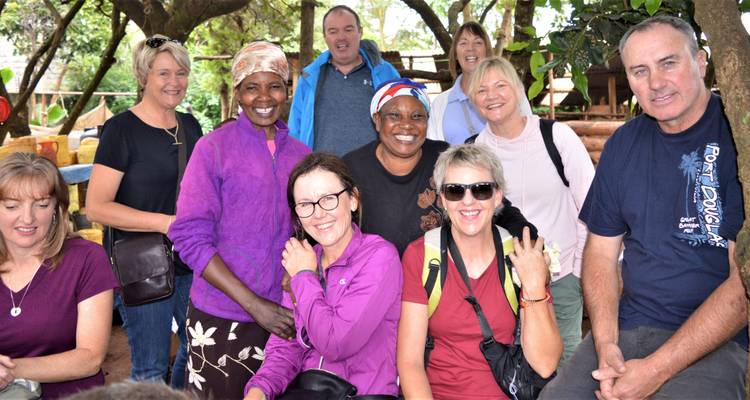 Un grupo de turistas sonrientes posa con mujeres locales bajo los árboles frondosos.