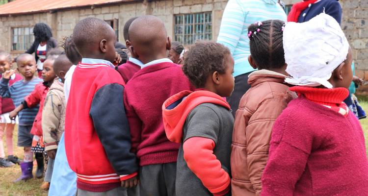 Niños pequeños con uniformes escolares alineados afuera de un edificio de aulas de ladrillo.