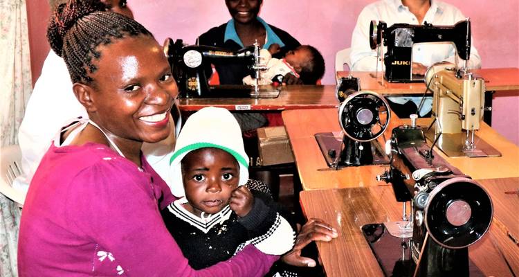 Mujer sonriente sosteniendo a un niño en una clase de costura con varias máquinas de coser de fondo.