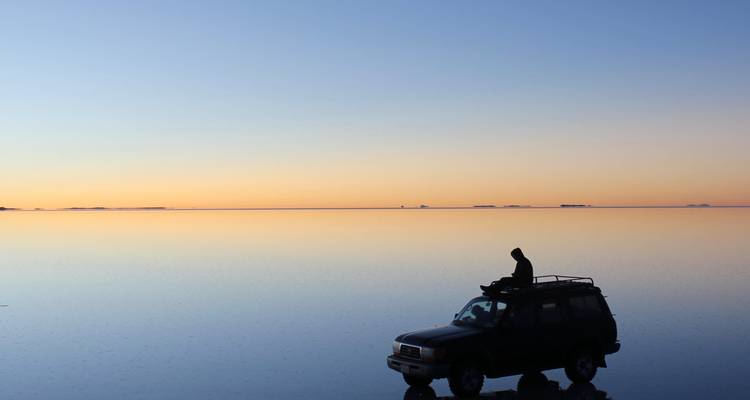 Silhouette d'un voyageur au sommet d'un 4x4 sur la surface miroir du Salar de Uyuni au coucher du soleil.