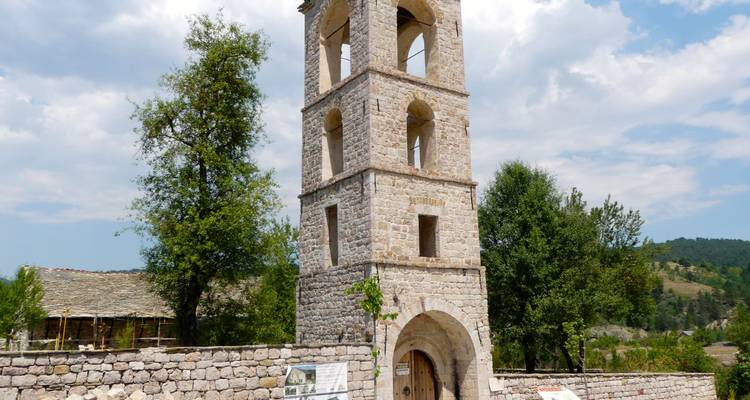 A tall stone bell tower rises above a low wall and countryside greenery under patchy clouds.