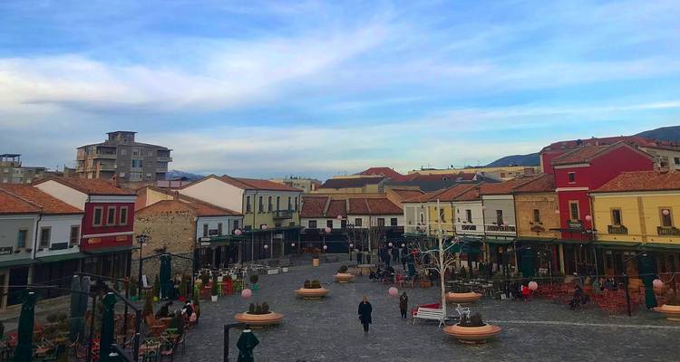 A spacious cobblestone plaza framed by colorful low buildings and cafés in an Albanian town on a clear day.