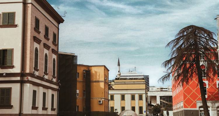 Colorful facades and a mosque minaret rise against a textured sky in a Balkan cityscape.