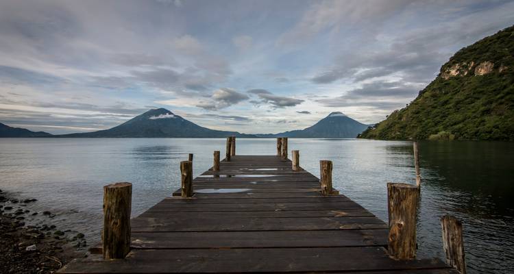 Un muelle de madera que se adentra en las aguas tranquilas del Lago Atitlán con dos volcanes imponentes en el horizonte