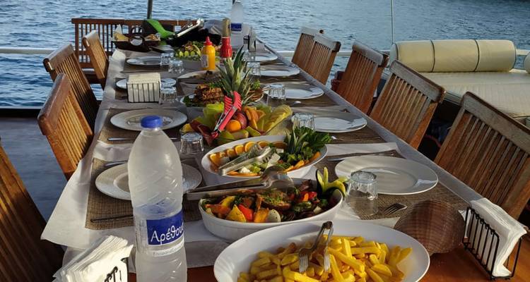 Tisch zum Essen im Freien auf dem Deck mit mediterranen Speisen, während das Schiff in ruhigen Gewässern vor Anker liegt.