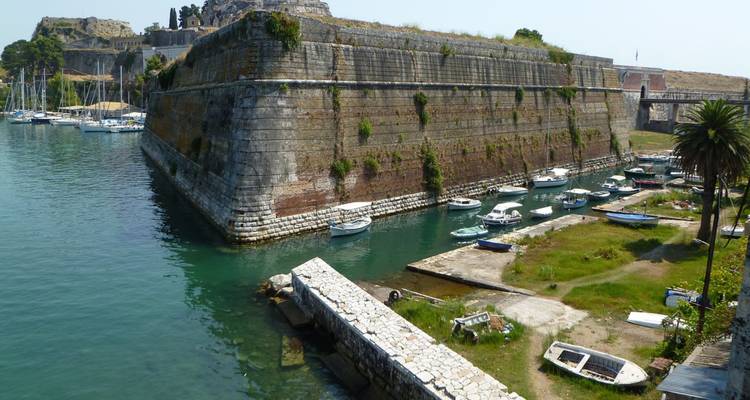 Massive Steinbefestigungsmauern führen hinab zu einem kleinen Hafen mit vertäuten Booten in Korfu.