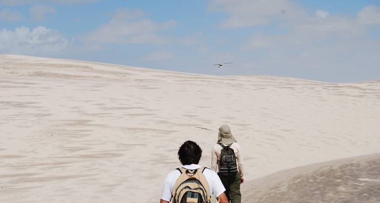Deux voyageurs en sac à dos traversent de vastes dunes de sable blanc sous un ciel bleu aux Lençóis Maranhenses