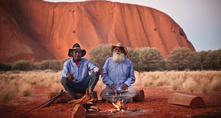 Zwei Aboriginal-Älteste sitzen an einem kleinen Feuer, während sich Ulurusgewaltige rote Felswand hinter ihnen im warmen Abendlicht erhebt.