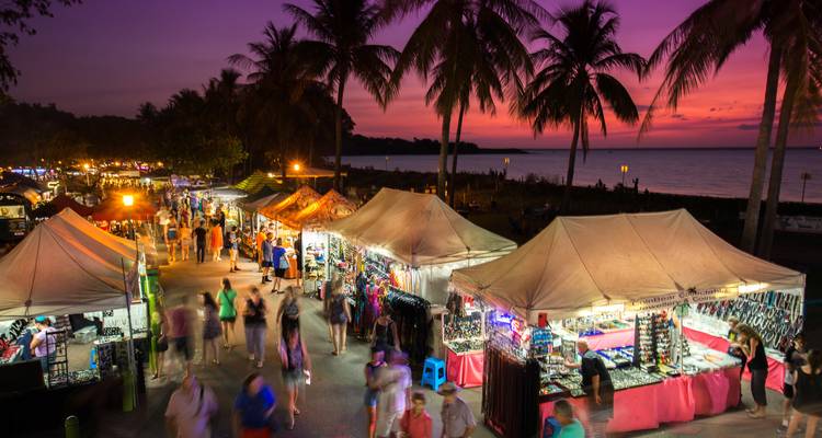 Geschäftiger Nachtmarkt am Meer mit farbenfrohen Ständen unter Palmen und einem magentafarbenen Sonnenuntergangshimmel.