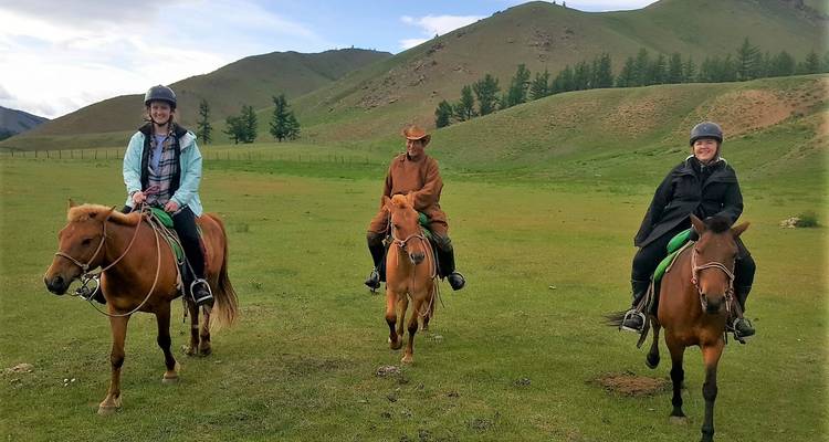 Drie ruiters op stoere Mongoolse paarden trekken door een open groen dal met zachte heuvels.