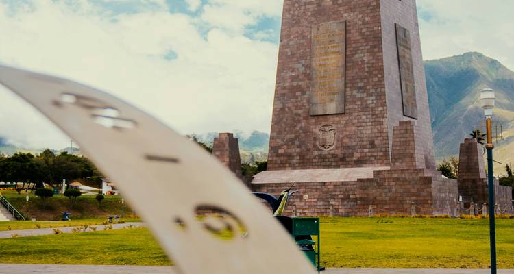 Das Äquatordenkmal "Mitad del Mundo" erhebt sich vor einer bergigen Kulisse an einem teilweise bewölkten Tag.
