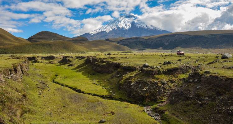 Breites Grastal mit erodierten Rinnen, die zum entfernten, schneebedeckten Cotopaxi unter verstreuten Wolken führen.