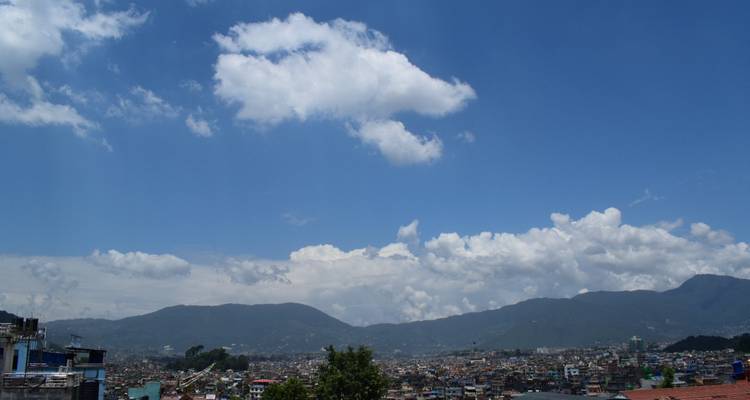Breite Tagesansicht über die Stadt Kathmandu mit fernen grünen Hügeln und verstreuten weißen Wolken gegen einen hellen blauen Himmel.