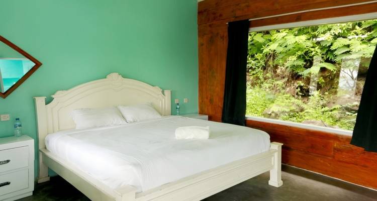 Habitación simple con pared verde menta, cama blanca y una gran ventana con vista al bosque.