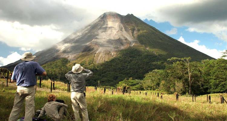 Los excursionistas observan el cono imponente de un volcán activo elevándose sobre las laderas boscosas bajo cielos parcialmente nublados.