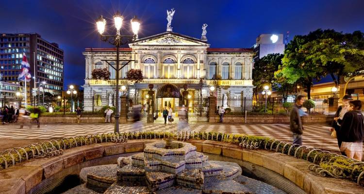 Teatro neoclásico iluminado que brilla en una animada plaza de la ciudad por la noche con una fuente de diseño en primer plano.