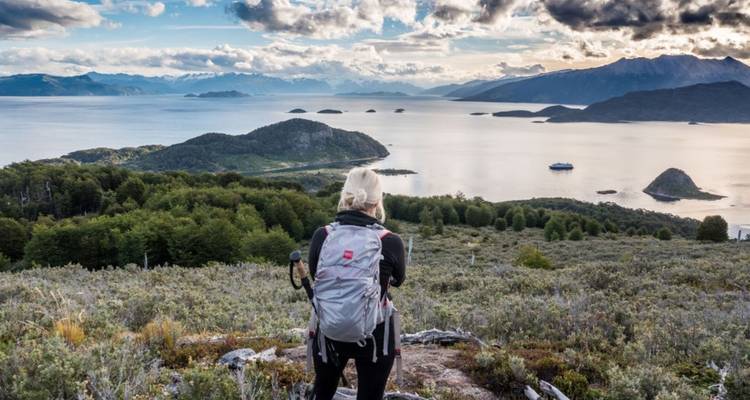 Ein Rucksacktourist blickt auf den mit Inseln gespickten Beagle-Kanal von einem hohen Grat unter dramatischen Wolken.