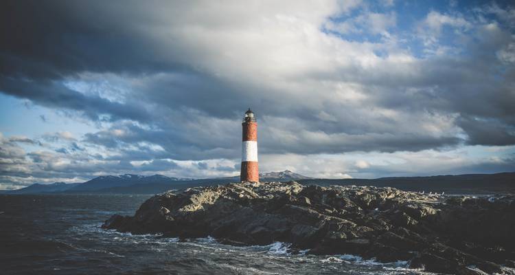 Der rot-weiß gestreifte Leuchtturm steht auf rauen Felsen vor einem stimmungsvollen patagonischen Himmel.