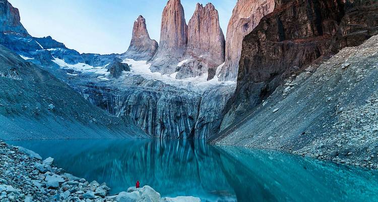 Person in red jacket stands by a mirror-like turquoise lake beneath the towering granite spires of Torres del Paine