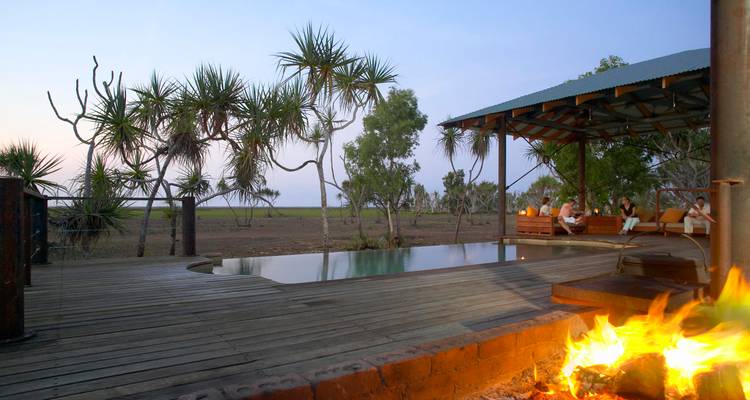 Abgelegenes Öko-Lodge-Deck mit Infinity-Pool, Feuerstelle und Schraubenbäumen mit Blick auf offene Feuchtgebiete in der Abenddämmerung.