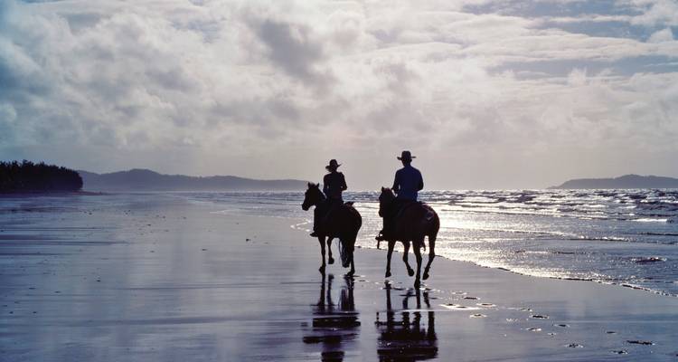Silhouettierte Reiter zu Pferd machen sich ihren Weg entlang eines spiegelnden, windgepeitschten Strandes unter einem bewölkten Himmel.
