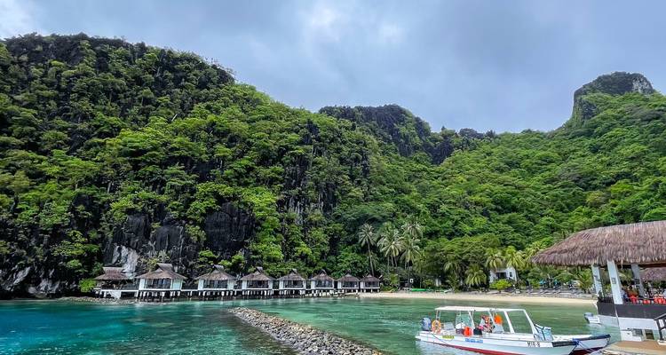Overwater villa's liggen langs een private baai met smaragdgroen water met daarachter imposante kalksteenrotsen in El Nido, terwijl een speedboat bezoekers naar de kust brengt.