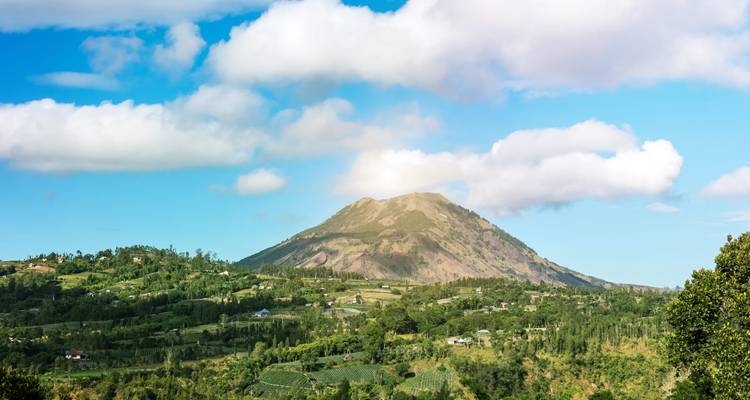 Vallée verdoyante avec des fermes dispersées sous un volcan solitaire, sous un ciel bleu éclatant et des nuages cotonneux.