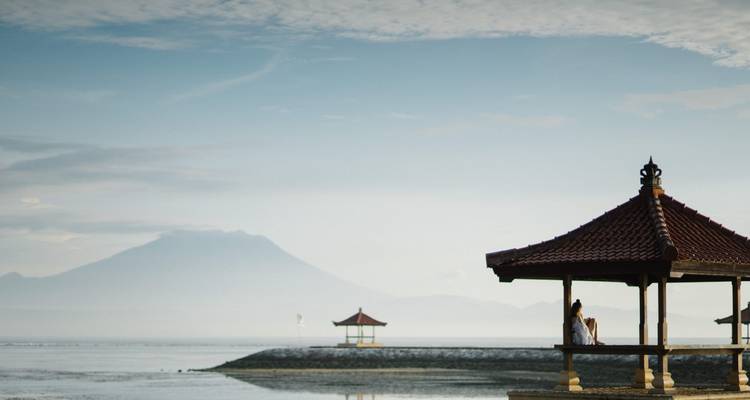 Pavillon balinais traditionnel sur un front de mer calme avec un volcan lointain s'élevant à travers la brume matinale.