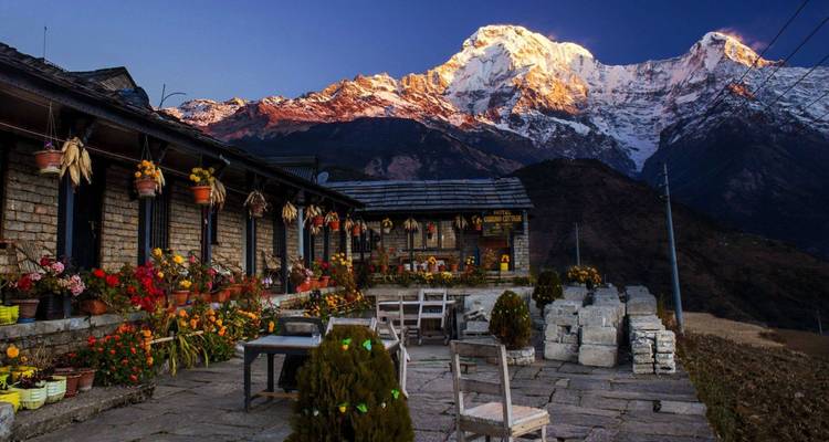 Flower-lined stone courtyard of a mountain lodge faces glowing snow peaks at dawn.