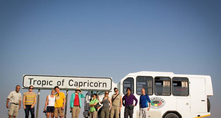 Groupe de touristes posant à côté d'un panneau routier du Tropique du Capricorne près d'un véhicule de safari sous un ciel bleu dégagé.