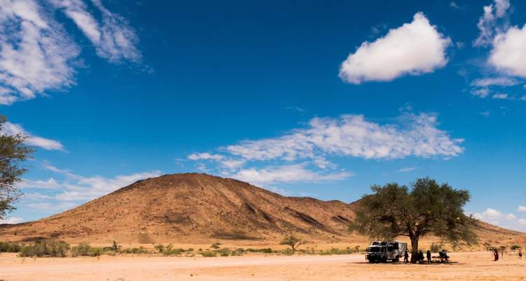 Vaste paysage désertique avec une colline basse, véhicule tout-terrain garé sous un acacia et voyageurs se reposant à proximité sous un ciel bleu éclatant parsemé de nuages épars.