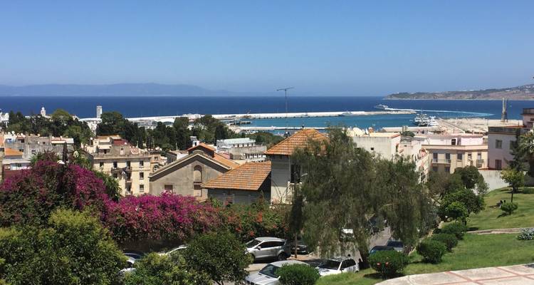 Vue panoramique sur les quartiers en pente de Tanger, la marina et la Méditerranée d'un bleu profond sous un ciel dégagé.