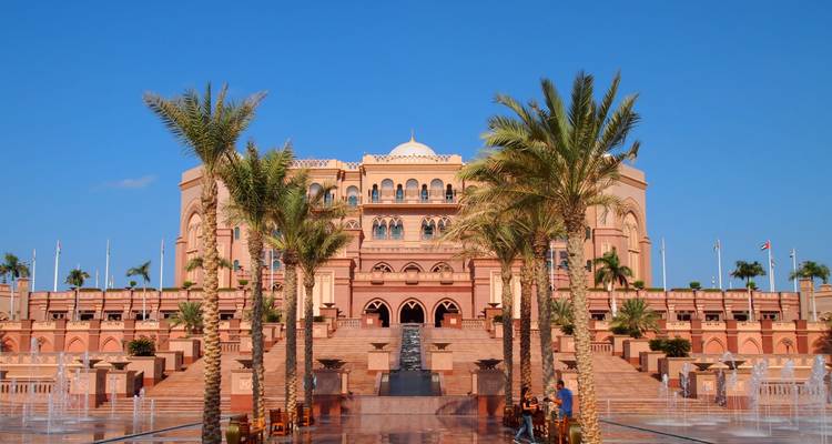 Gran fachada del Emirates Palace enmarcada por palmeras y fuentes bajo un cielo azul despejado de Abu Dabi.