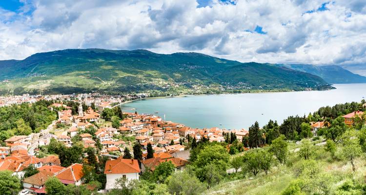 Vista panorámica amplia del Lago Ohrid con el pueblo de techos de terracota abajo, rodeado de montañas verde exuberante bajo nubes dramáticas.