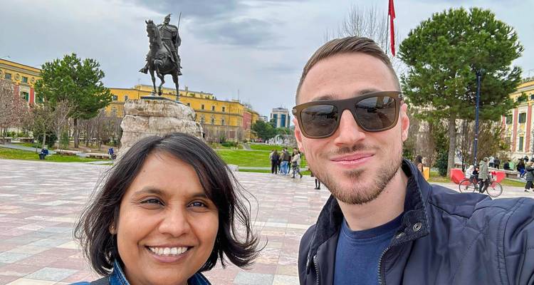 Alegre selfie de dos viajeros con gafas de sol en la Plaza Skanderbeg con la estatua y los edificios coloridos detrás de ellos.